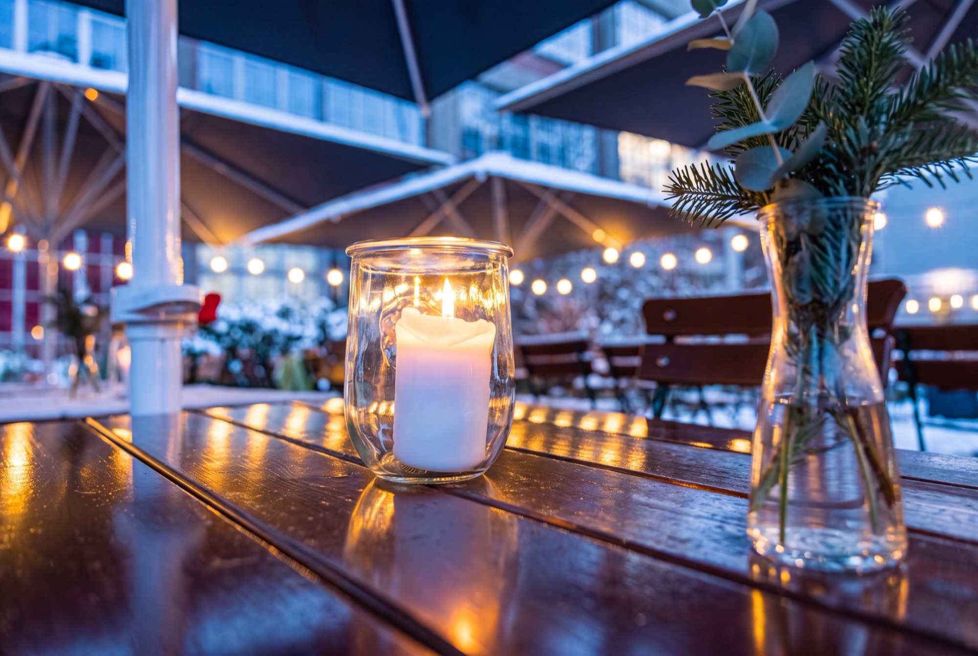Kerze im Windglas auf einem Holztisch der Terrasse, dahinter Lichterketten und Schnee in der blauen Stunde