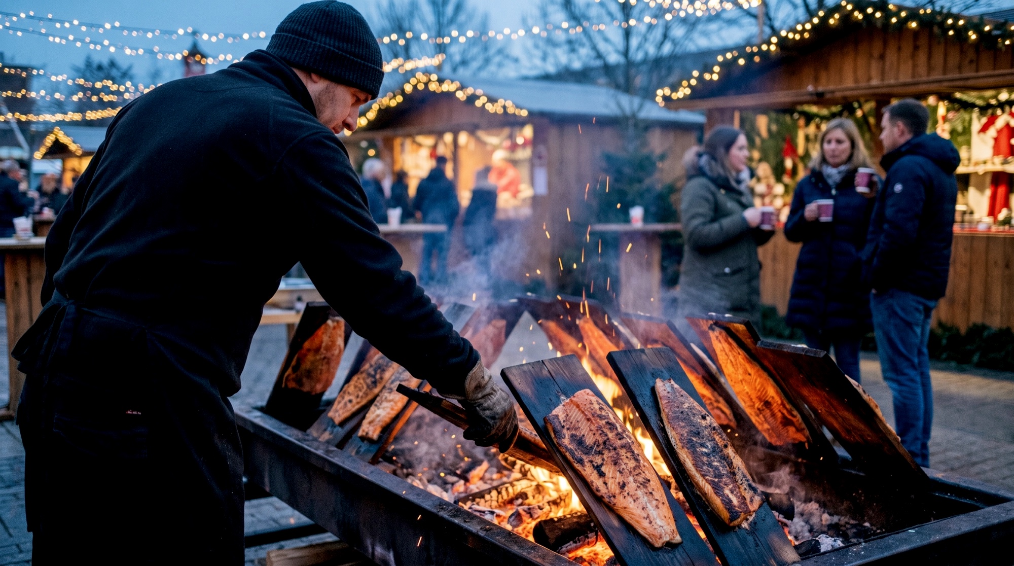 Firmenweihnachtsmarkt: Flammlachsbretter mit Lachsseiten &uuml;ber der offenen Glut, Mitarbeiter in Schwarz richtet ein Brett aus, Buden mit Lichterketten in der blauen Stunde