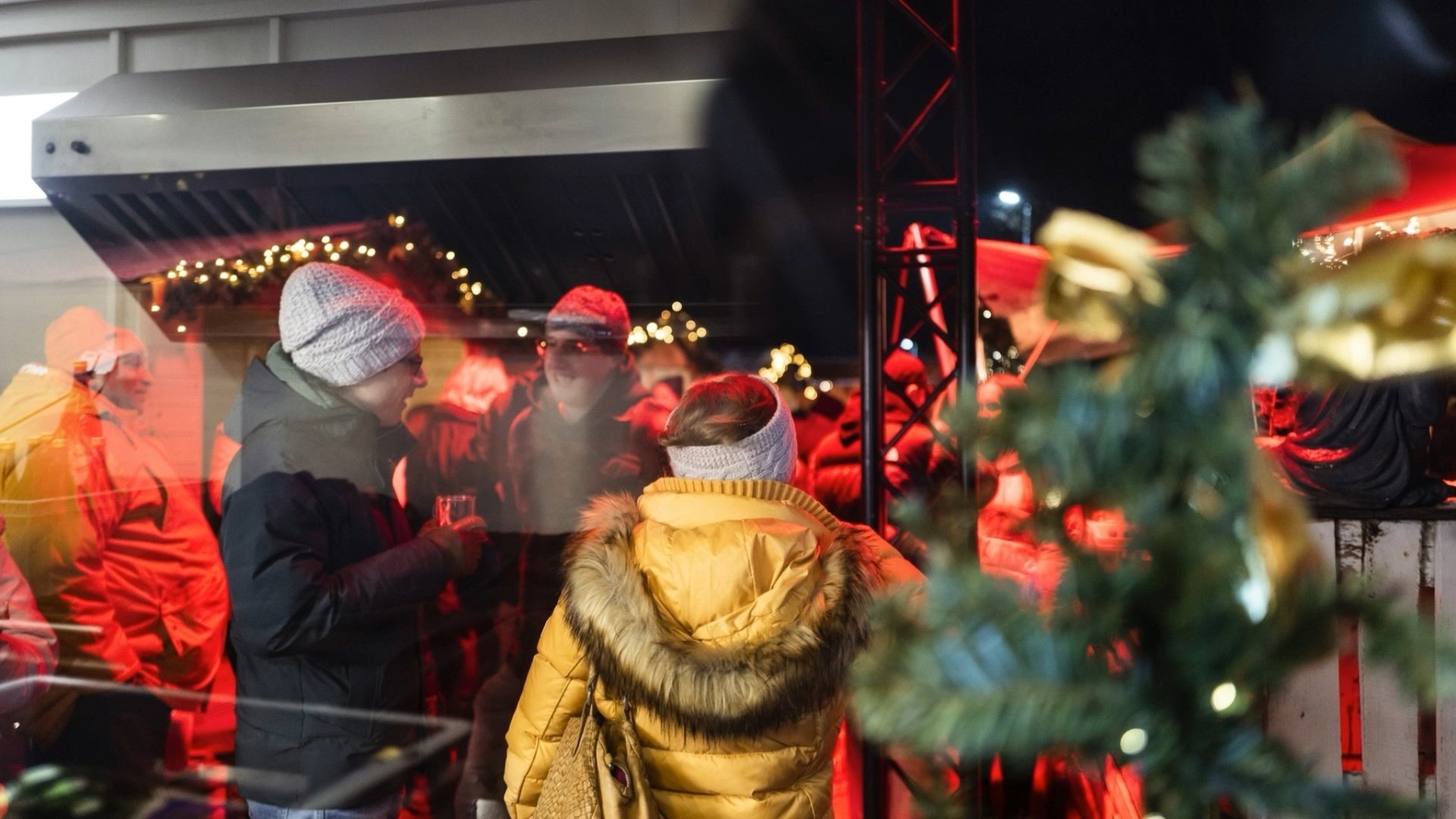 G&auml;ste auf einem Firmen-Weihnachtsmarkt am Abend, vorn ein geschm&uuml;ckter Tannenzweig mit Lichterkette